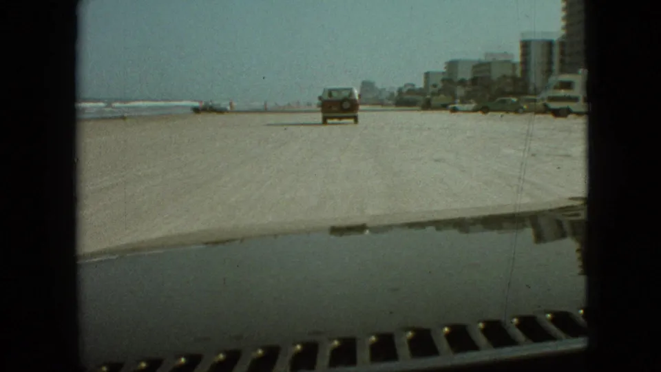 The moving camera follows a station wagon across a sandy beach-like roadway, with water at left, parked cars at right, and mid-century buildings lining the background.