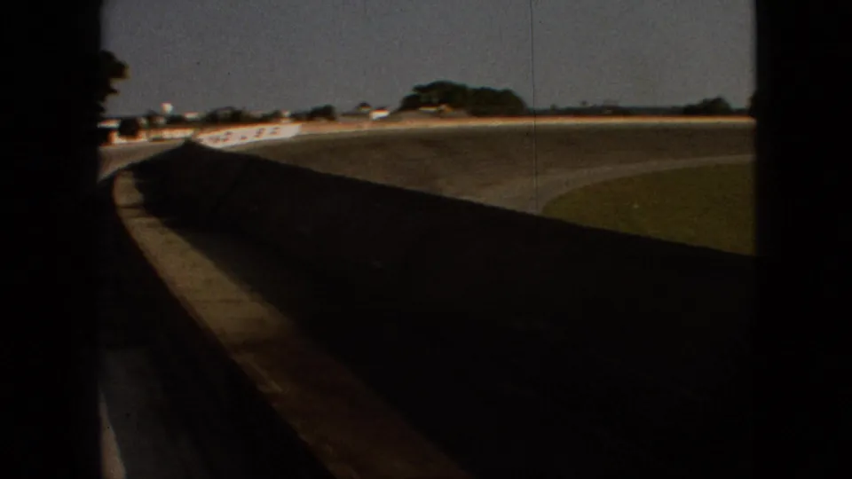 The clip shifts to an in-vehicle view of a curved concrete bridge or overpass, with a dark road edge, grassy area, and small vehicles visible in the distance.