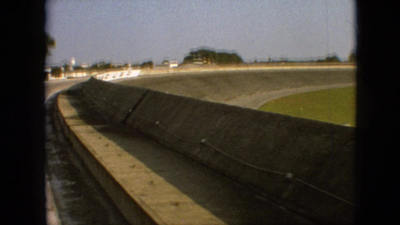 1968 Branson, Missouri 8mm home-movie frame showing a first-person view from inside a car as it follows a banked racetrack curve beside a concrete retaining wall and grassy infield.