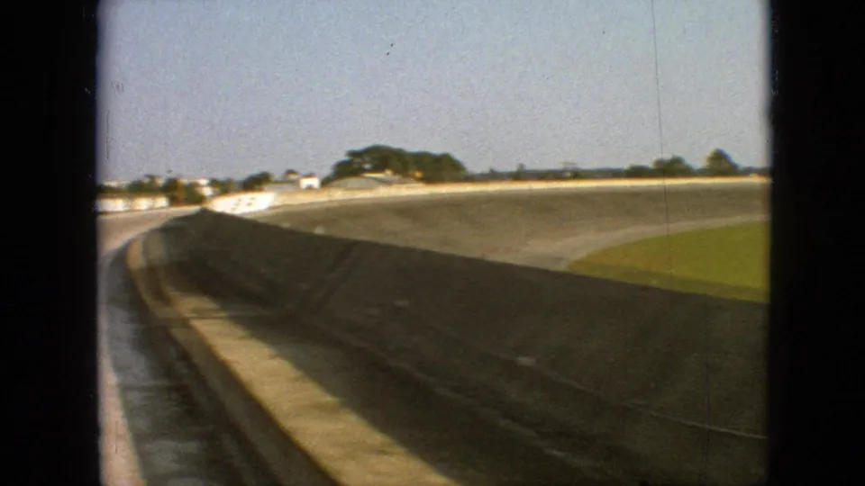 1968 Branson, Missouri 8mm home-movie frame showing a car-window-framed view of a banked track curve, concrete wall, grassy infield, and distant tree line.