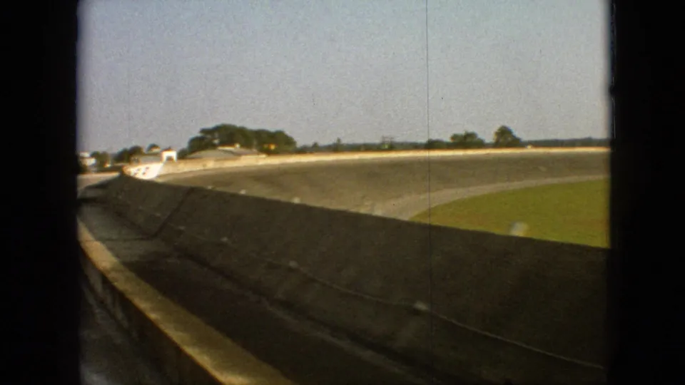 1968 Branson, Missouri 8mm frame showing the passenger perspective beside a concrete retaining wall as the track curves around a grassy infield under a pale sky.