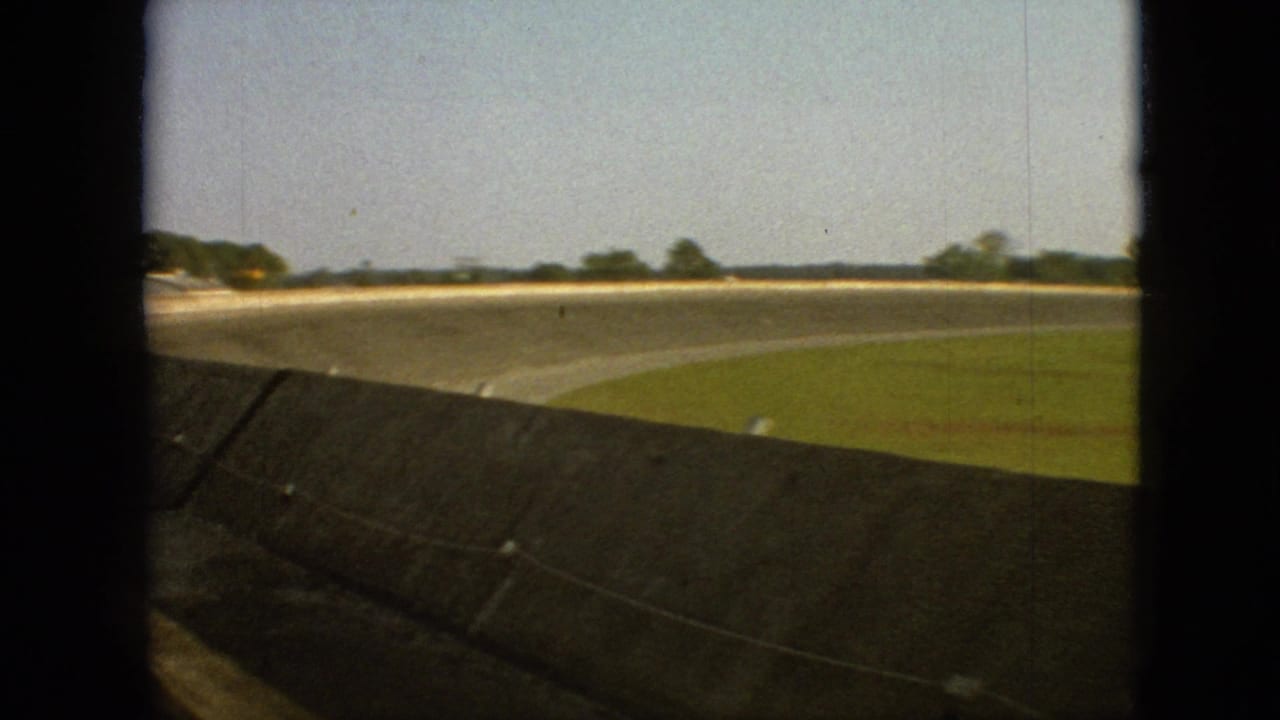 1968 Branson, Missouri home-movie frame showing a curved highway from a moving automobile, with a concrete barrier, grass, open sky, and distant trees.