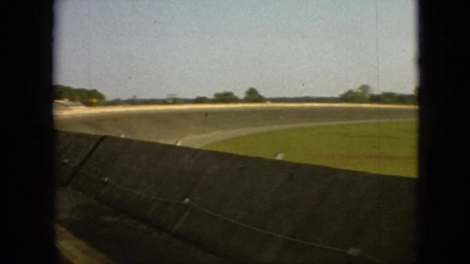 1968 Branson, Missouri home-movie frame showing a curved highway from a moving automobile, with a concrete barrier, grass, open sky, and distant trees.