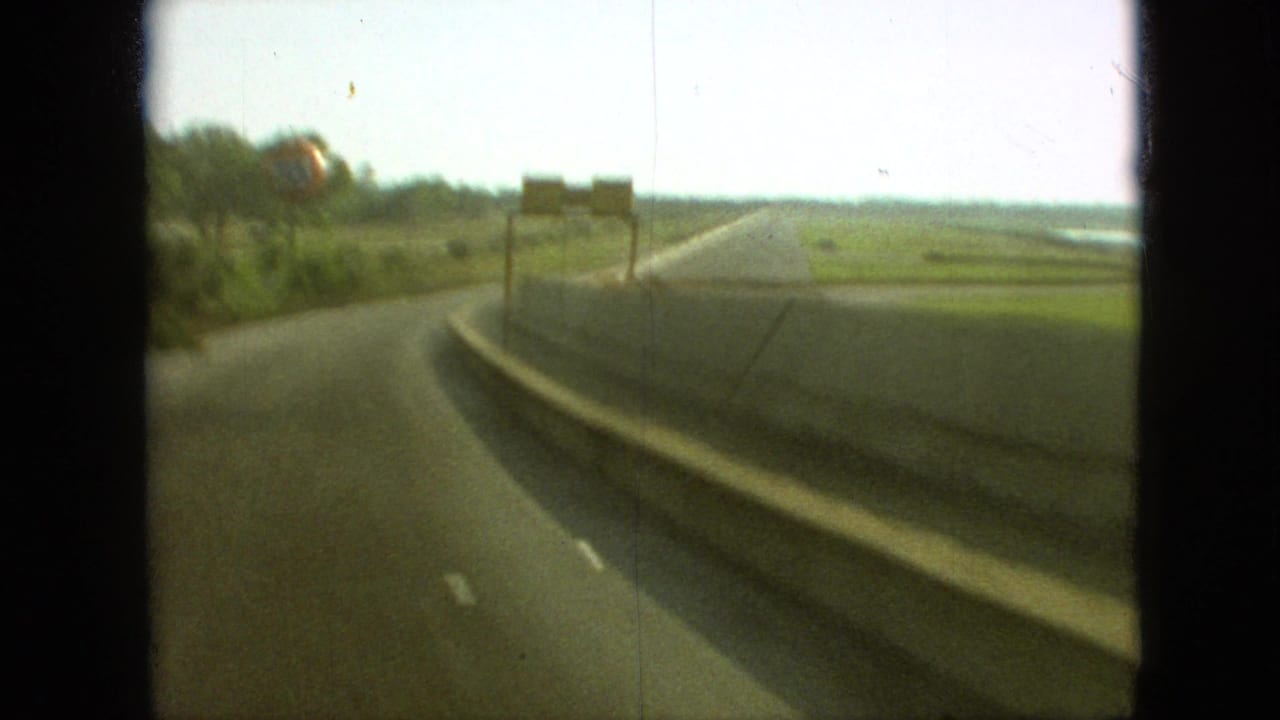 Roadway curves beside open fields with roadside signs ahead and dark film borders.
