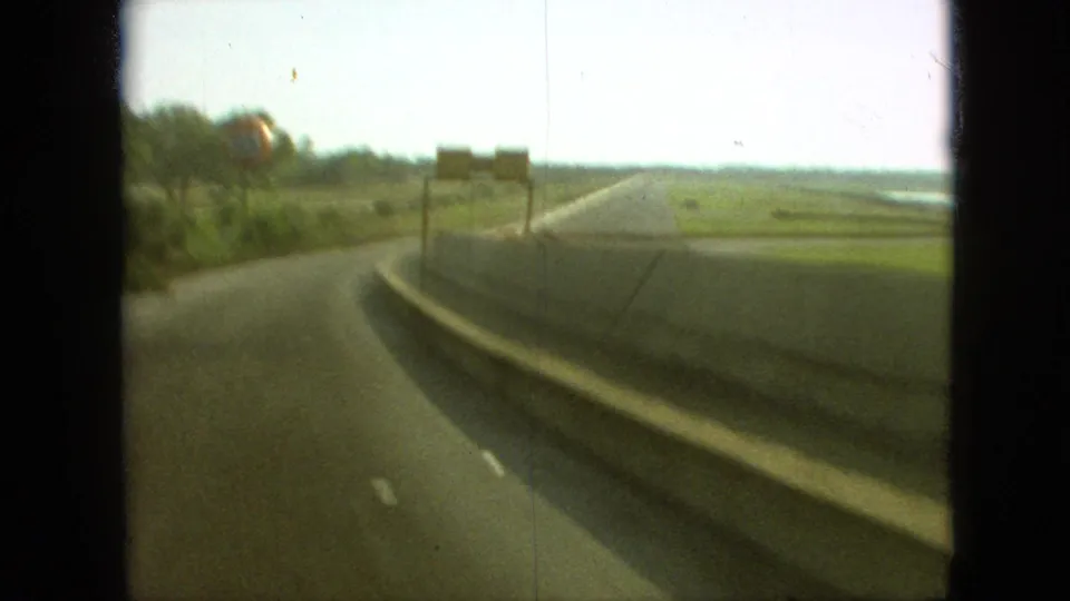 First-person automobile view on a Branson, Missouri highway in 1968, looking toward a road curve, open fields, distant signs, and a bright sky.