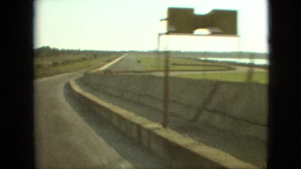 1968 Branson, Missouri road-travel frame with a blurred sign in the foreground, a curving highway, concrete barrier, and flat open fields beyond.