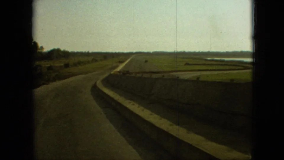 Moving automobile view in 1968 Branson, Missouri showing the highway bend continuing past concrete barriers, open grassland, and a distant tree line.