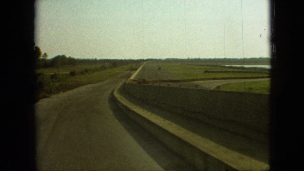 1968 Branson, Missouri home-movie frame showing a paved rural road curving past fields, a concrete roadside barrier, distant trees, and dark car-window edges.