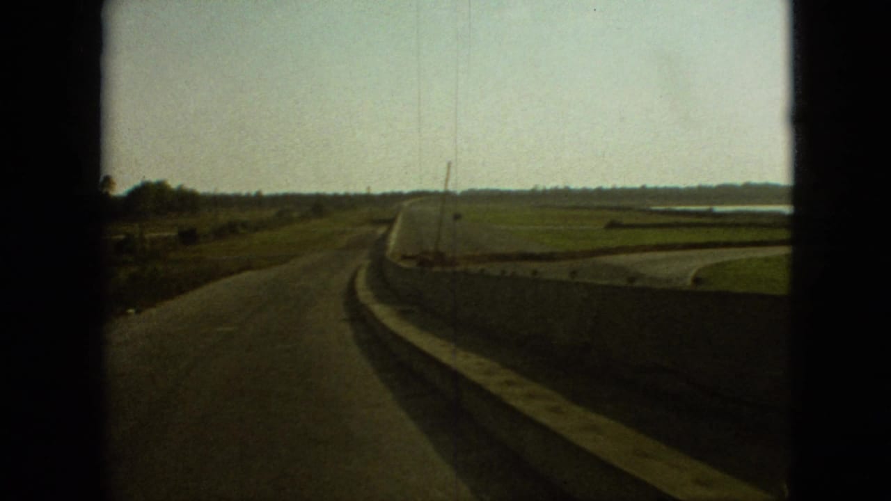 Rural highway bends ahead with fields, roadside barrier, and distant trees.