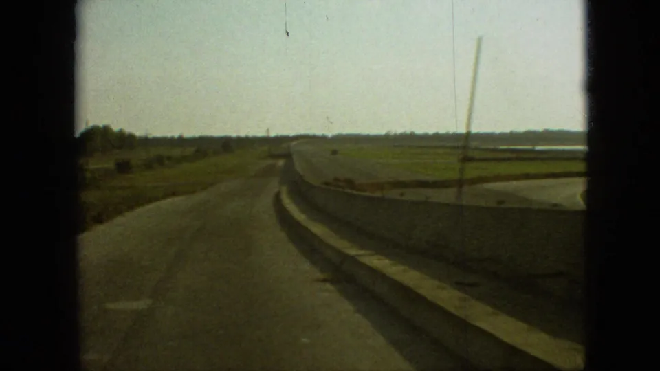 1968 Branson, Missouri frame from a moving car showing the road surface, long concrete barrier, open fields, distant trees, and visible film scratches.