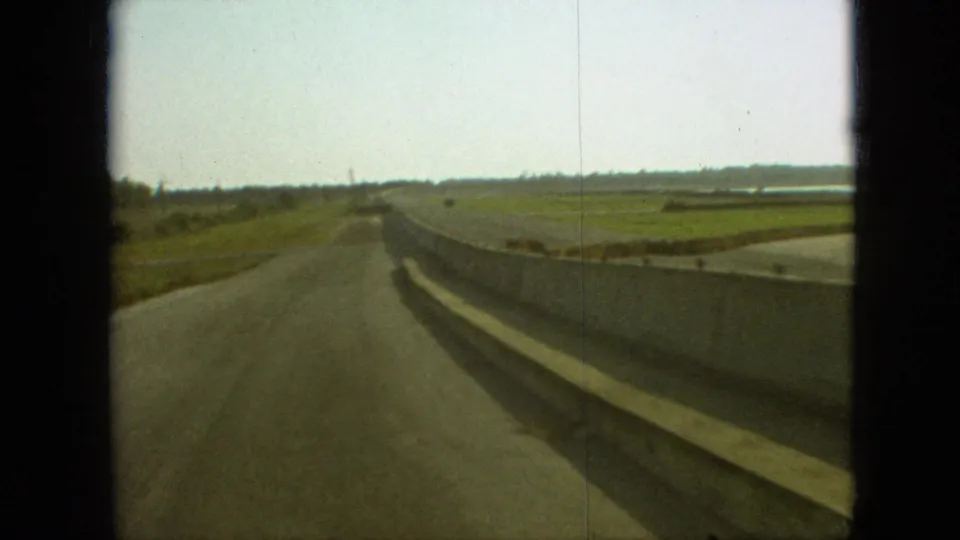 1968 Branson, Missouri home-movie frame showing a rural road extending ahead beside a concrete barrier, with green fields and distant trees under a bright sky.
