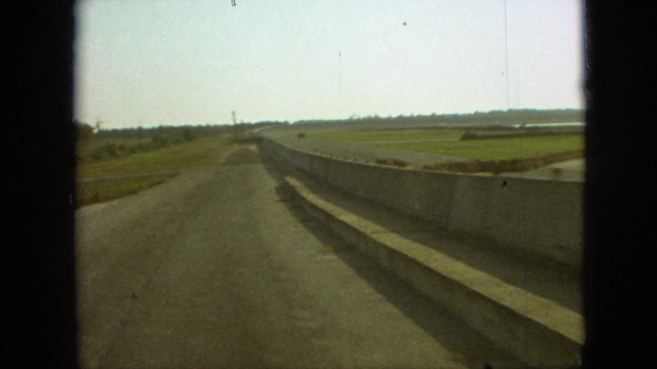 1968 8mm home-movie view from a truck window near Branson, Missouri, showing a rural road, concrete barrier, green farmland, and pale sky.