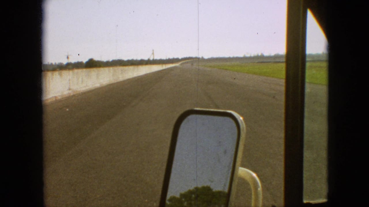 Straight highway view with a side mirror and concrete barrier beside farmland.