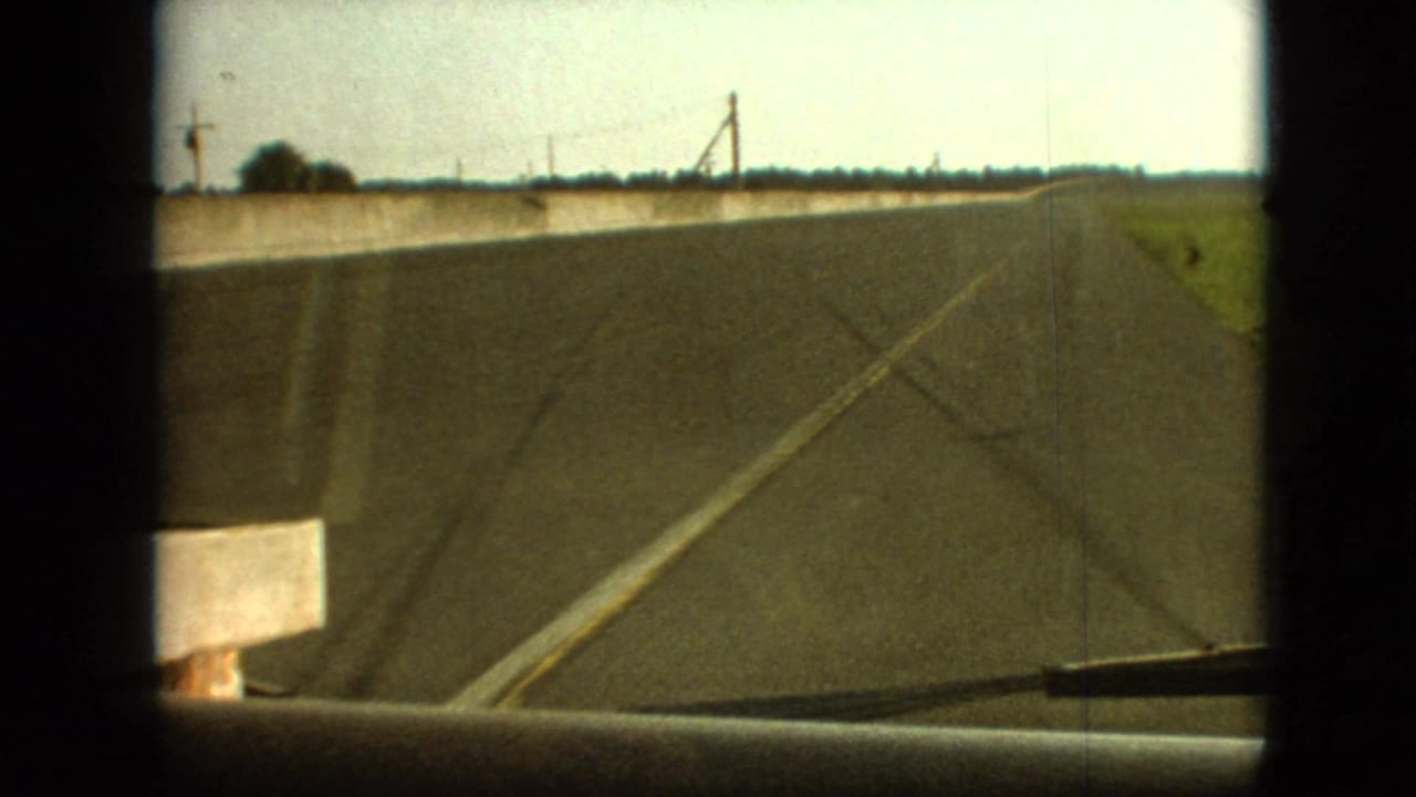 1968 8mm home-movie frame from the Branson, Missouri area showing a forward vehicle view along a straight rural road bordered by fields and a low concrete barrier.