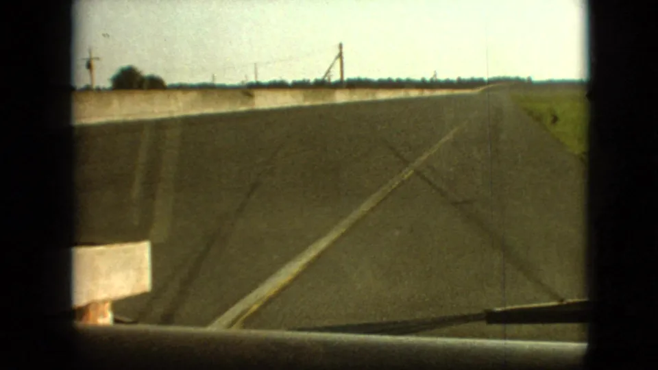 1968 8mm home-movie frame from the Branson, Missouri area showing a forward vehicle view along a straight rural road bordered by fields and a low concrete barrier.