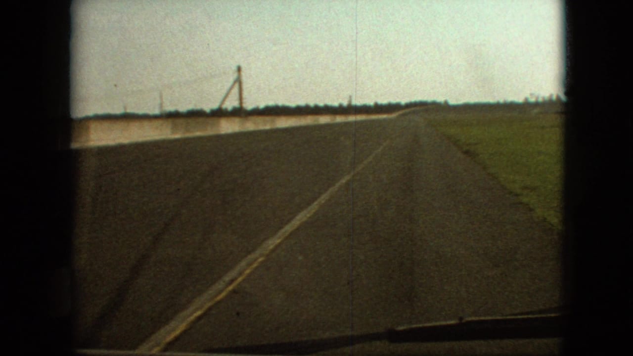 Forward-facing 1968 8mm frame showing a long rural road near Branson, Missouri, with open fields, a left-side barrier, utility poles, and soft faded color.