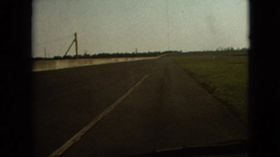 1968 Branson-area home-movie frame showing a moving-vehicle perspective along a quiet country road with open grassland and sparse roadside utility structures.