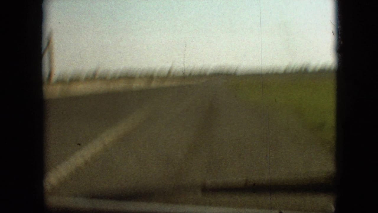 1968 8mm home-movie frame from the Branson, Missouri area showing a blurred forward view of a curved rural road, grassy edge, pale sky, and dark film-frame borders.