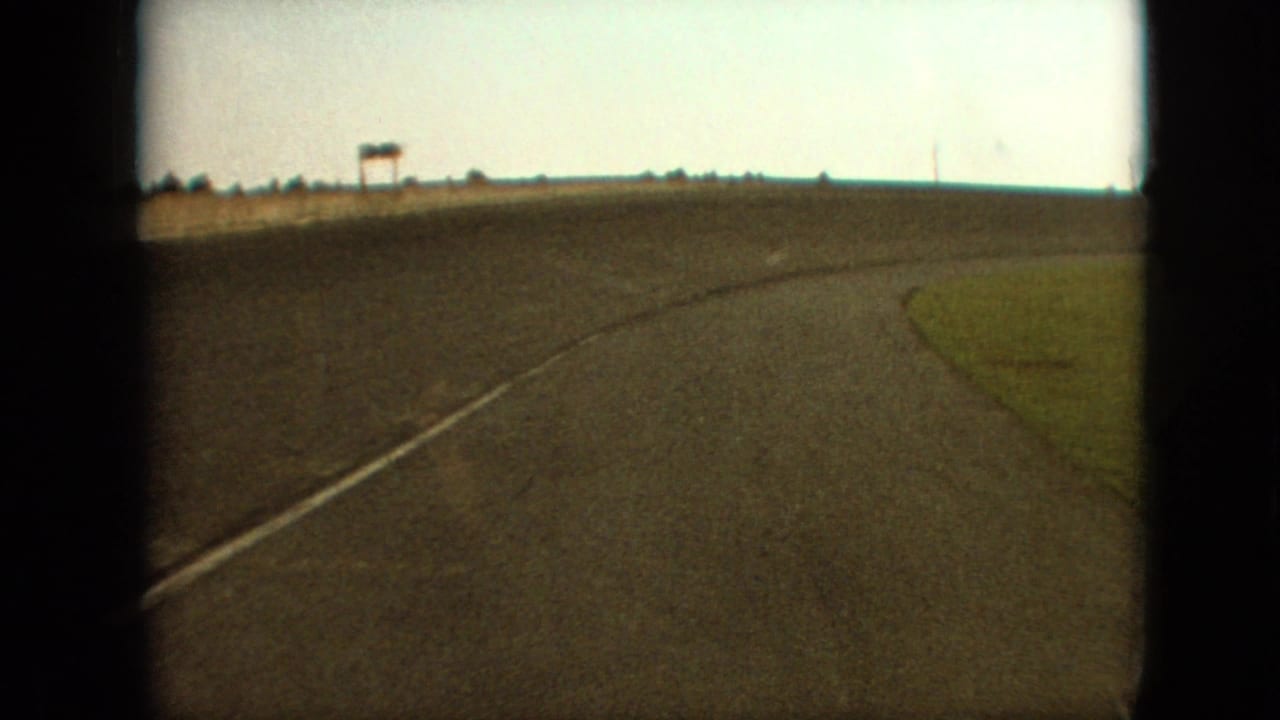 1968 Branson, Missouri area road view from a moving vehicle, with a right-curving asphalt surface, grassy shoulder, distant sign-like structure, and pale sky.