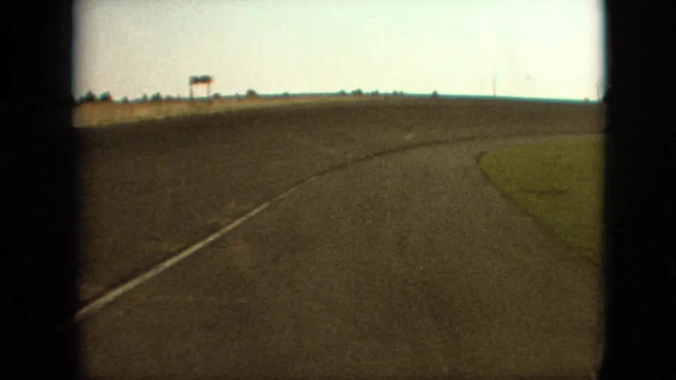 1968 Branson, Missouri area road view from a moving vehicle, with a right-curving asphalt surface, grassy shoulder, distant sign-like structure, and pale sky.