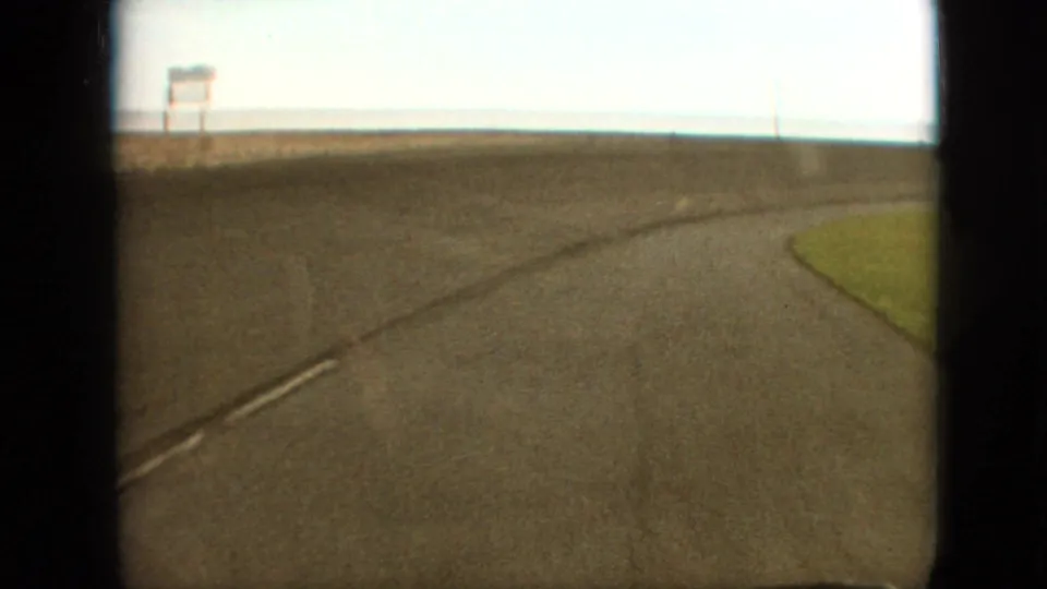 1968 8mm road-trip frame near Branson, Missouri showing a paved curve, painted center or edge line, grassy shoulder, low horizon, and distant roadside sign-like structure.