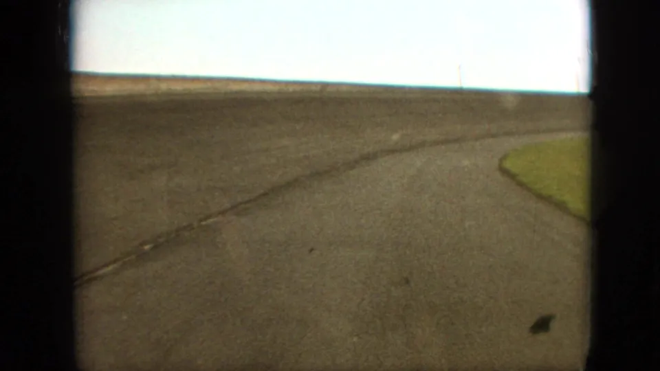 1968 Branson, Missouri area home-movie frame showing the moving-vehicle view of a rural asphalt road bending right, with grass, open horizon, a utility pole, and 8mm grain.