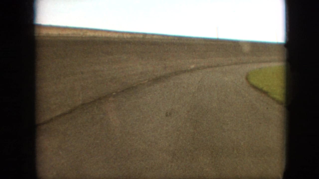 1968 Branson, Missouri home-movie frame showing a low moving view of a curving asphalt race track with a small grassy infield edge and heavy dark gate borders.