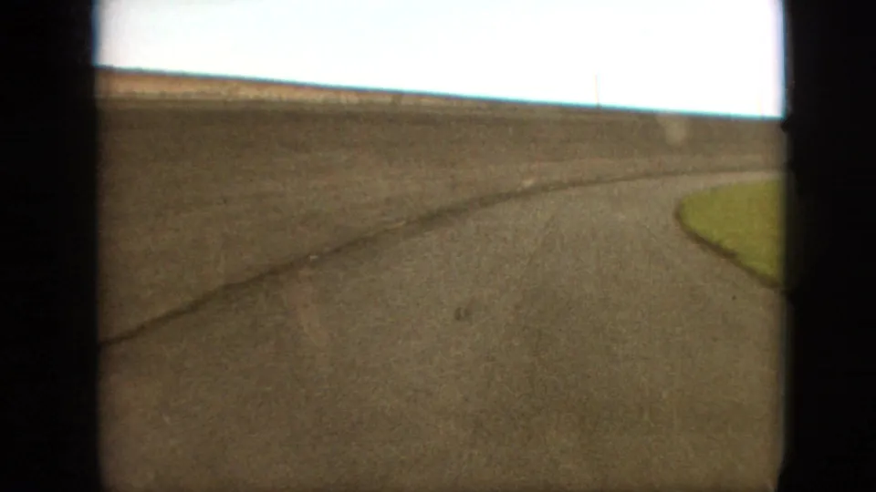 1968 Branson, Missouri home-movie frame showing a low moving view of a curving asphalt race track with a small grassy infield edge and heavy dark gate borders.