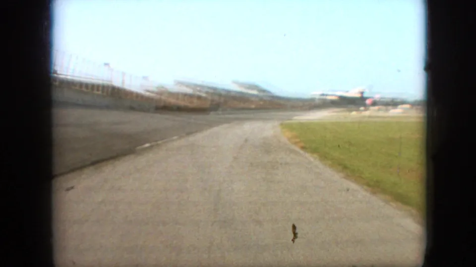 1968 Branson, Missouri frame showing the track opening toward a distant grandstand or spectator structure, with safety barriers, fencing, grass, and bright sky visible.