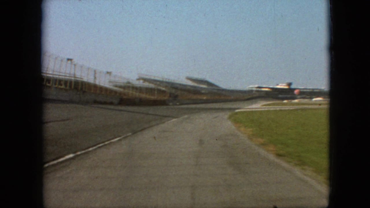 1968 Branson, Missouri home-movie frame showing a forward-moving driver perspective on an empty racetrack beside open grandstands and a grass verge.