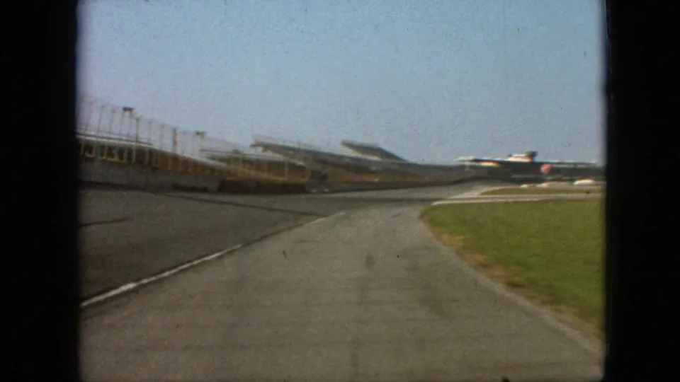 1968 Branson, Missouri home-movie frame showing a forward-moving driver perspective on an empty racetrack beside open grandstands and a grass verge.