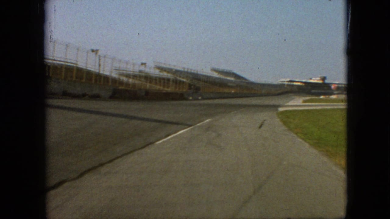 1968 Branson, Missouri frame from a moving vehicle on an empty racetrack, with grandstand structures to the left and grass bordering the track at right.