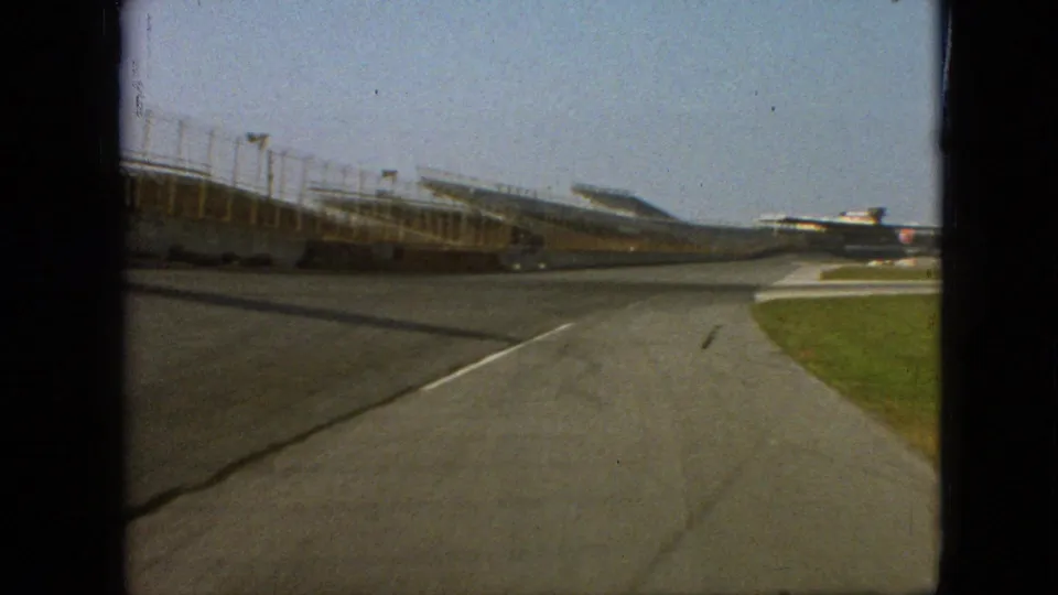 1968 Branson, Missouri frame from a moving vehicle on an empty racetrack, with grandstand structures to the left and grass bordering the track at right.
