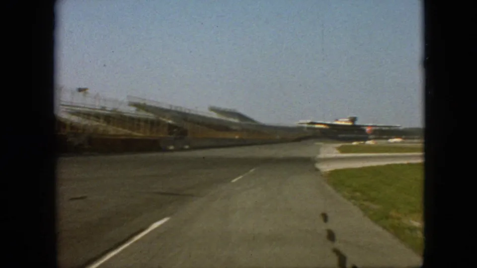 1968 Branson, Missouri home-movie still showing the vehicle perspective moving past empty racetrack seating, with white markings on the asphalt and grass along the right side.
