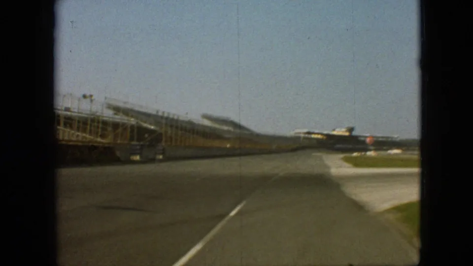 1968 Branson, Missouri frame showing an empty racetrack straightaway from a driver’s perspective, with grandstands to the left, grass at the edge, and distant structures ahead.