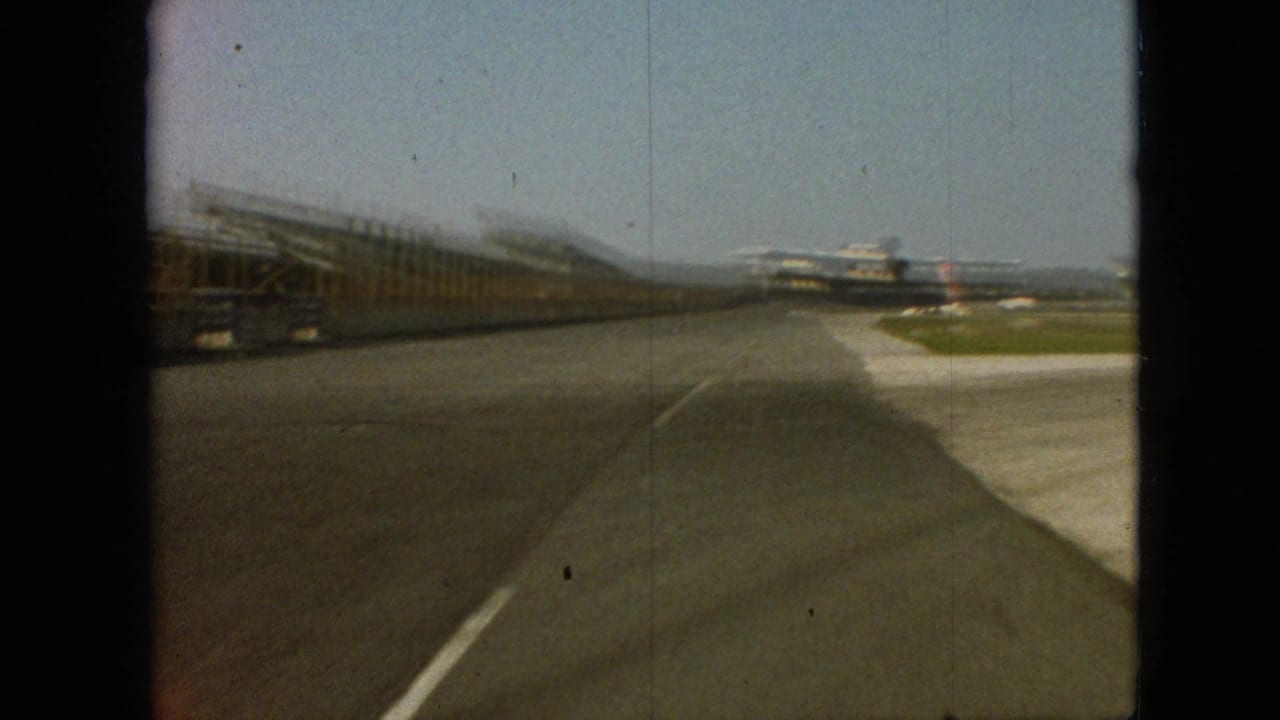 1968 Branson, Missouri 8mm frame showing a forward-moving view along a broad paved lane, with long low structures on the left and distant aircraft-like forms ahead.