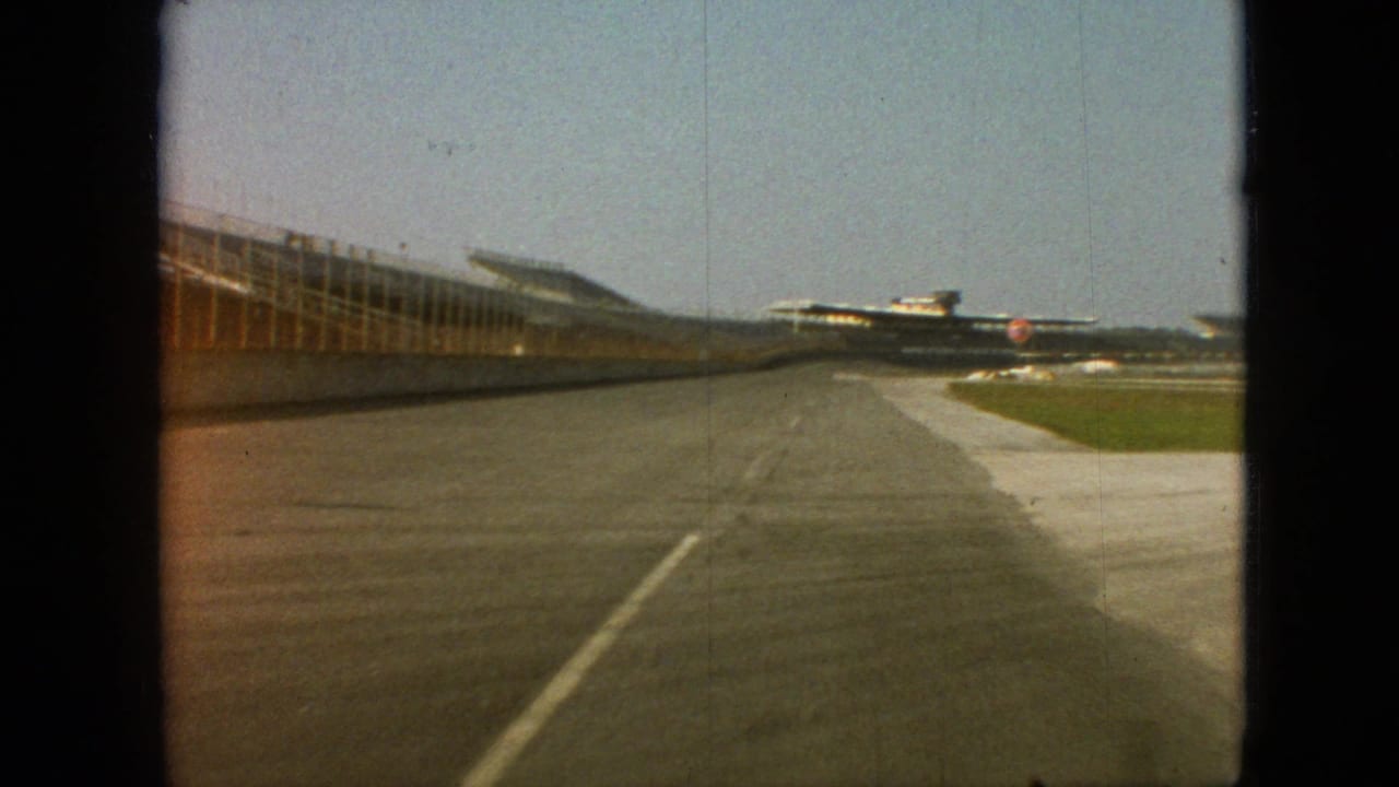 1968 Branson, Missouri 8mm frame with the camera moving along a paved lane beside a long structure, grass verge, and distant aircraft-like shapes.