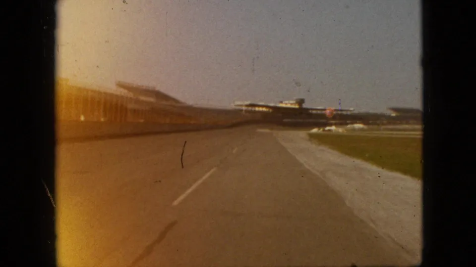 1968 Branson, Missouri 8mm frame with warm exposure flare over a paved lane, grassy edge, and distant aircraft-like forms near low structures.