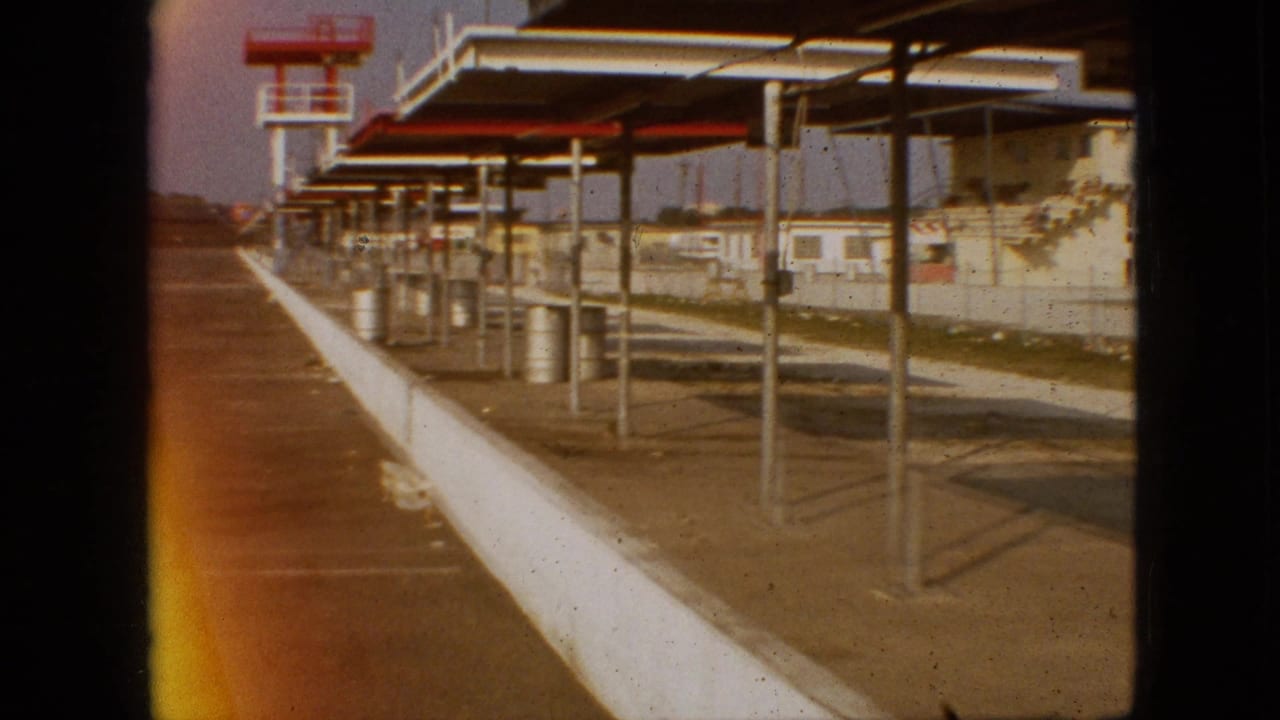 1968 Branson, Missouri 8mm frame showing a moving view along a raceway edge with a white concrete barrier, empty paved area, metal canopy posts, barrels, and low buildings beyond.