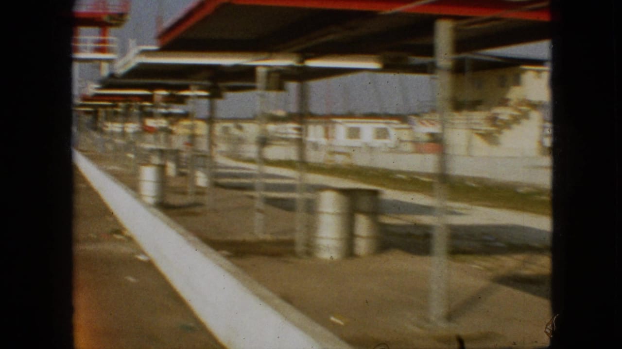 1968 Branson, Missouri frame from a moving 8mm view past trackside canopy structures, barrels, benches or platforms, and a white concrete barrier.