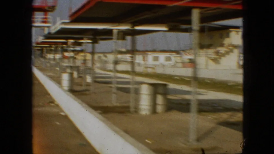 1968 Branson, Missouri frame from a moving 8mm view past trackside canopy structures, barrels, benches or platforms, and a white concrete barrier.