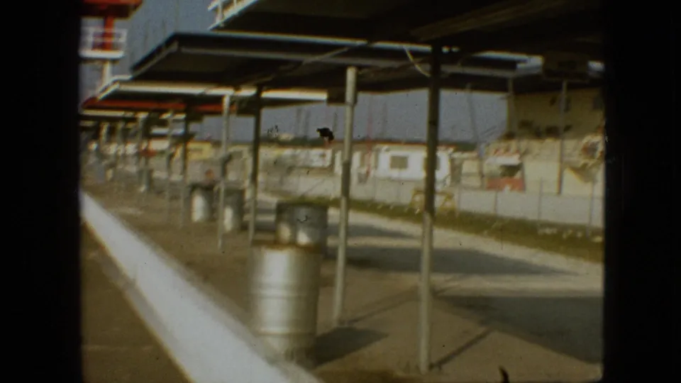 1968 Branson, Missouri 8mm frame with motion blur as the camera passes canopy supports, metal barrels, empty pavement, and a concrete barrier at a quiet raceway area.