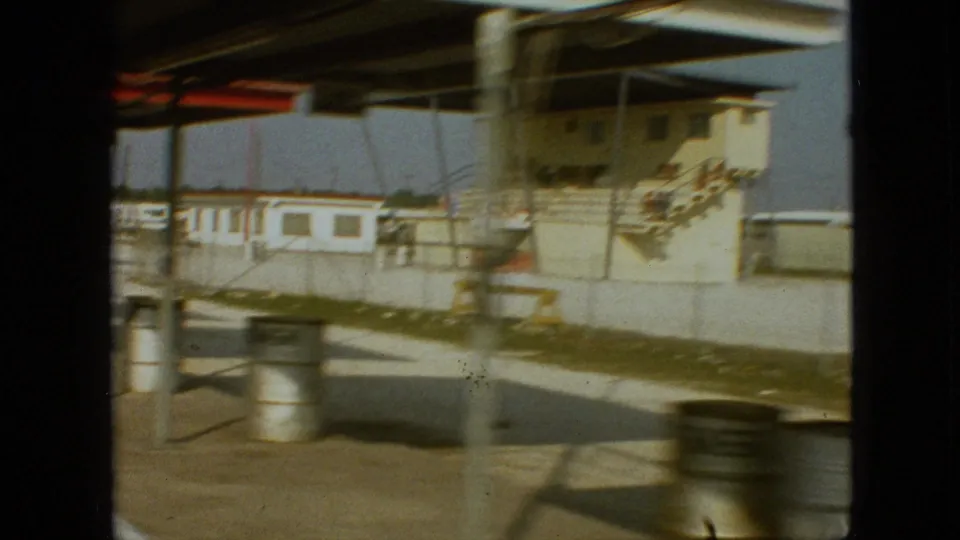 1968 Branson, Missouri frame showing a blurred close pass of metal barrels, canopy posts, pale building walls, red roof trim, and an empty paved raceway edge.