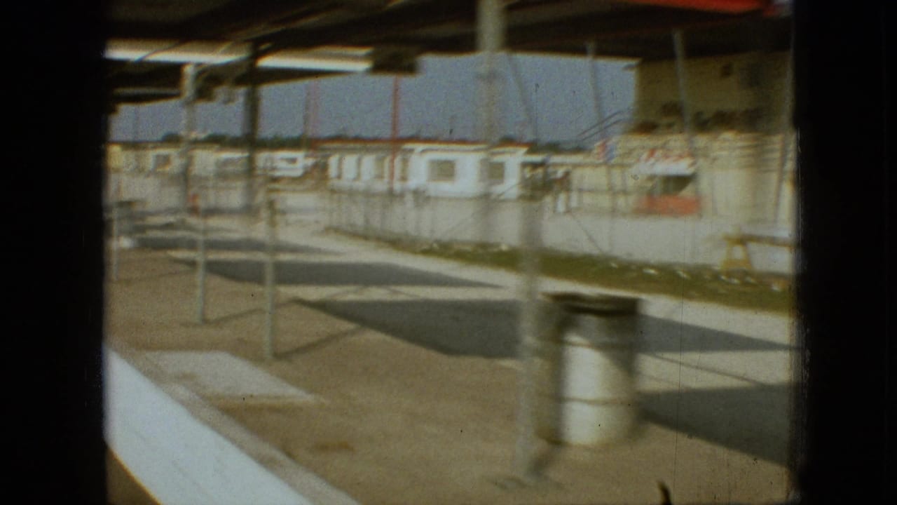 1968 Branson, Missouri home-movie frame from a moving vehicle showing a fenced walkway, low structures, metal posts, and dark vehicle-window framing near a stadium or racetrack complex.
