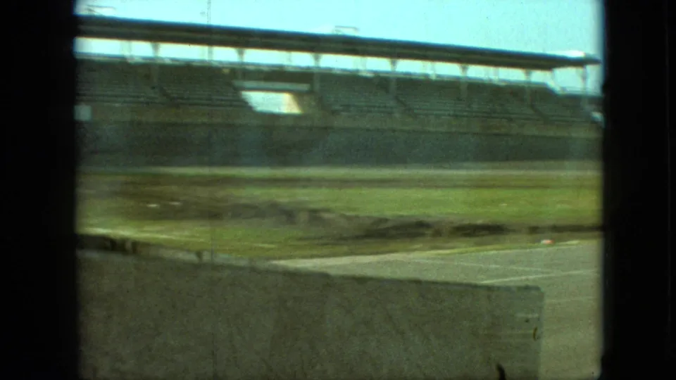 1968 Branson, Missouri home-movie frame showing empty stadium bleachers beyond a grassy infield and paved foreground, with vehicle-window framing and visible film grain.