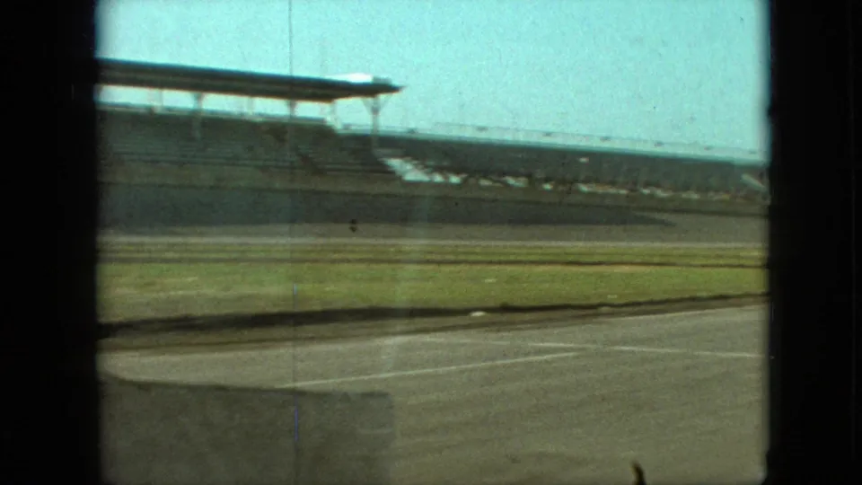 1968 Branson, Missouri frame with empty bleachers, a grassy field, paved lanes, and dark vehicle-window framing as the camera continues past the stadium or racetrack complex.