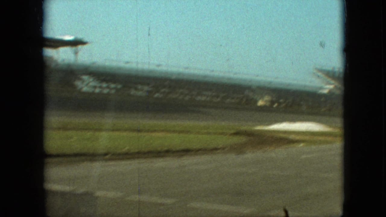 1968 Branson, Missouri 8mm frame showing an empty racetrack, grassy infield, and distant grandstand structures viewed from a moving vehicle.