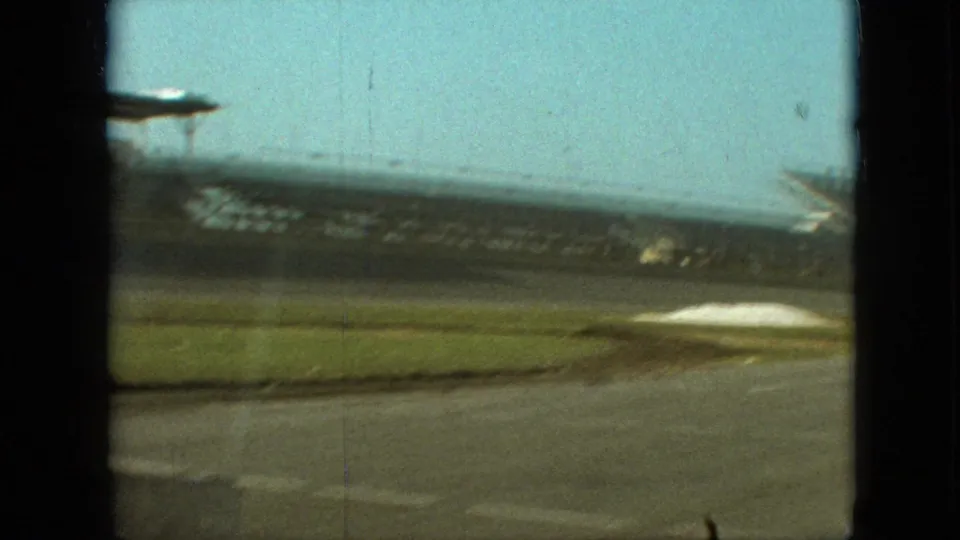 1968 Branson, Missouri 8mm frame showing an empty racetrack, grassy infield, and distant grandstand structures viewed from a moving vehicle.