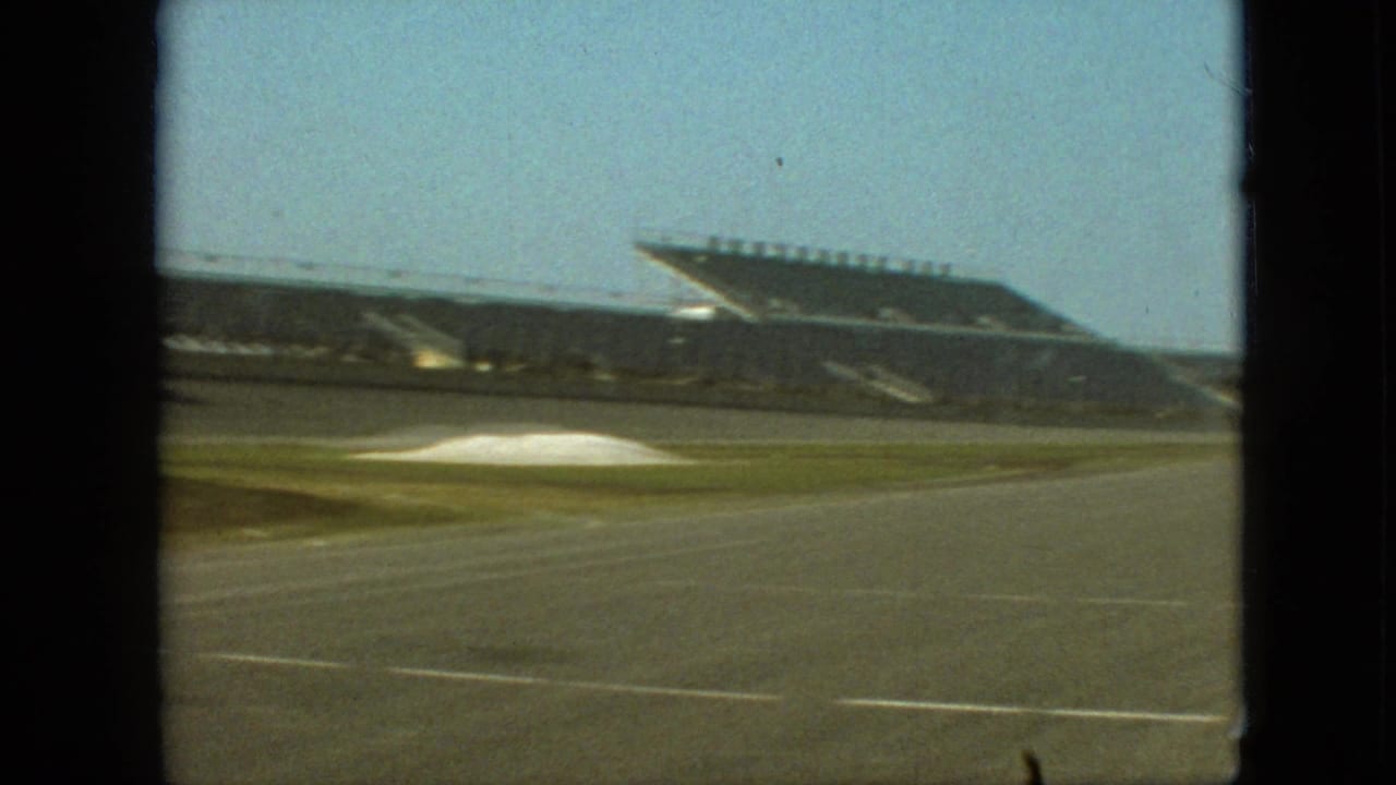 1968 Branson, Missouri racetrack view with a broad asphalt curve, grassy infield, and empty spectator stands in soft 8mm color.
