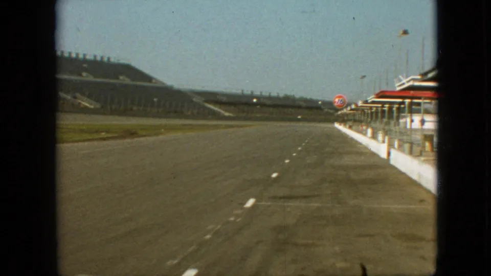 1968 Branson, Missouri 8mm frame looking down the pit lane past empty grandstands, red-roofed pit structures, barrels, and a visible racing-service brand mark requiring review.